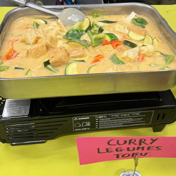 Un bac de curry de légumes au tofu sur son réchaud, avec son étiquette écrite à la main posée devant.