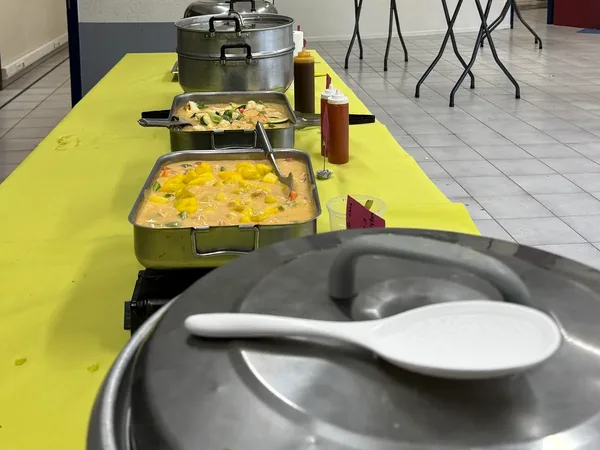 Une longue table de buffet nappée de jaune : paniers vapeur, bacs de curry et sauces, avant l'arrivée des invités ; au premier plan, le cuiseur à riz.